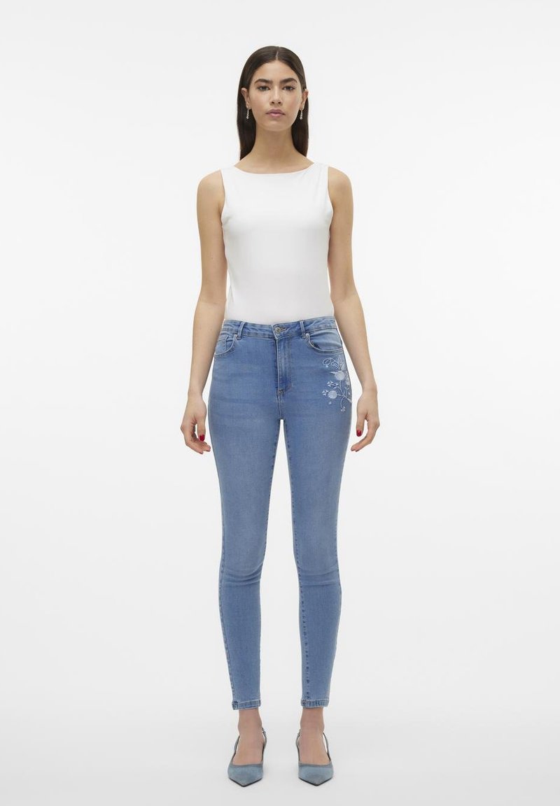 Skinny Jeans aus hellblauem Denim mit floralem Stickdetails am Oberschenkel, getragen mit einem weißen, figurbetonten Tanktop und hellblauen Absatzschuhen.