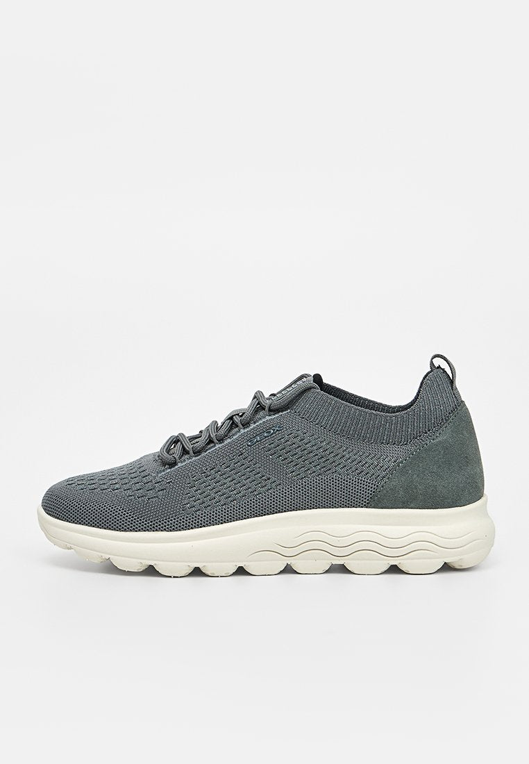 Grauer gestrickter Sneaker mit weißer welliger Sohle, vorderer Schnürung und Wildleder-Fersenpatch, von der Seite auf weißem Hintergrund gezeigt.