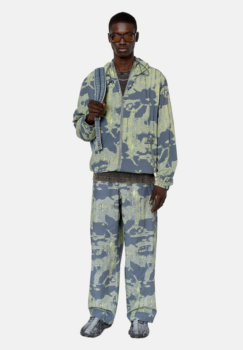Grau und gelb camouflierter Trainingsanzug, bestehend aus einer jacke mit Reißverschluss und lockeren Hosen. Zubehör umfasst einen gemusterten Gurt und eine Sonnenbrille.