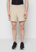 Mann, der beige Shorts, schwarze Sportschuhe und ein schwarzes ärmelloses Oberteil trägt, steht vor einem schlichten, hellen Hintergrund.