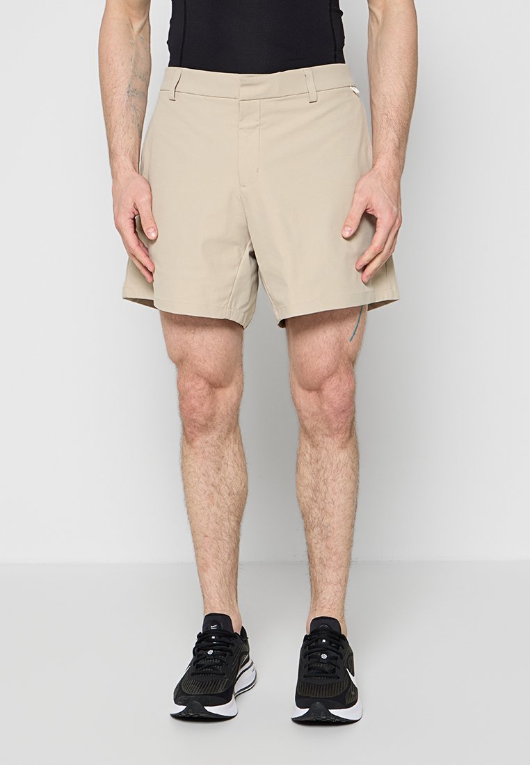 Mann, der beige Shorts, schwarze Sportschuhe und ein schwarzes ärmelloses Oberteil trägt, steht vor einem schlichten, hellen Hintergrund.
