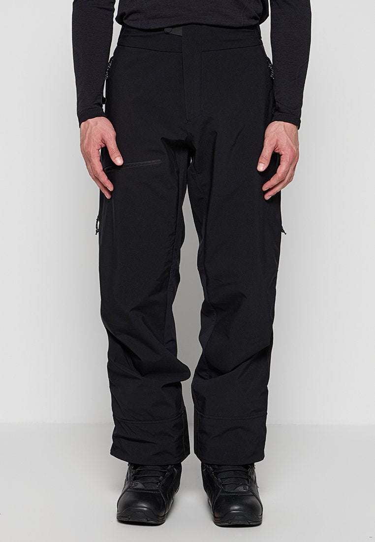 Mann trägt schwarze wasserdichte Hose mit Reißverschlusstaschen und schwarze Wanderschuhe, steht vor einem einfarbigen Hintergrund.