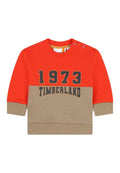 Roter und heller Pullover mit langen Ärmeln, rundem Ausschnitt und Knopfdetails an der Schulter. Mit der Aufschrift "1973 TIMBERLAND" in dunkler Schrift.