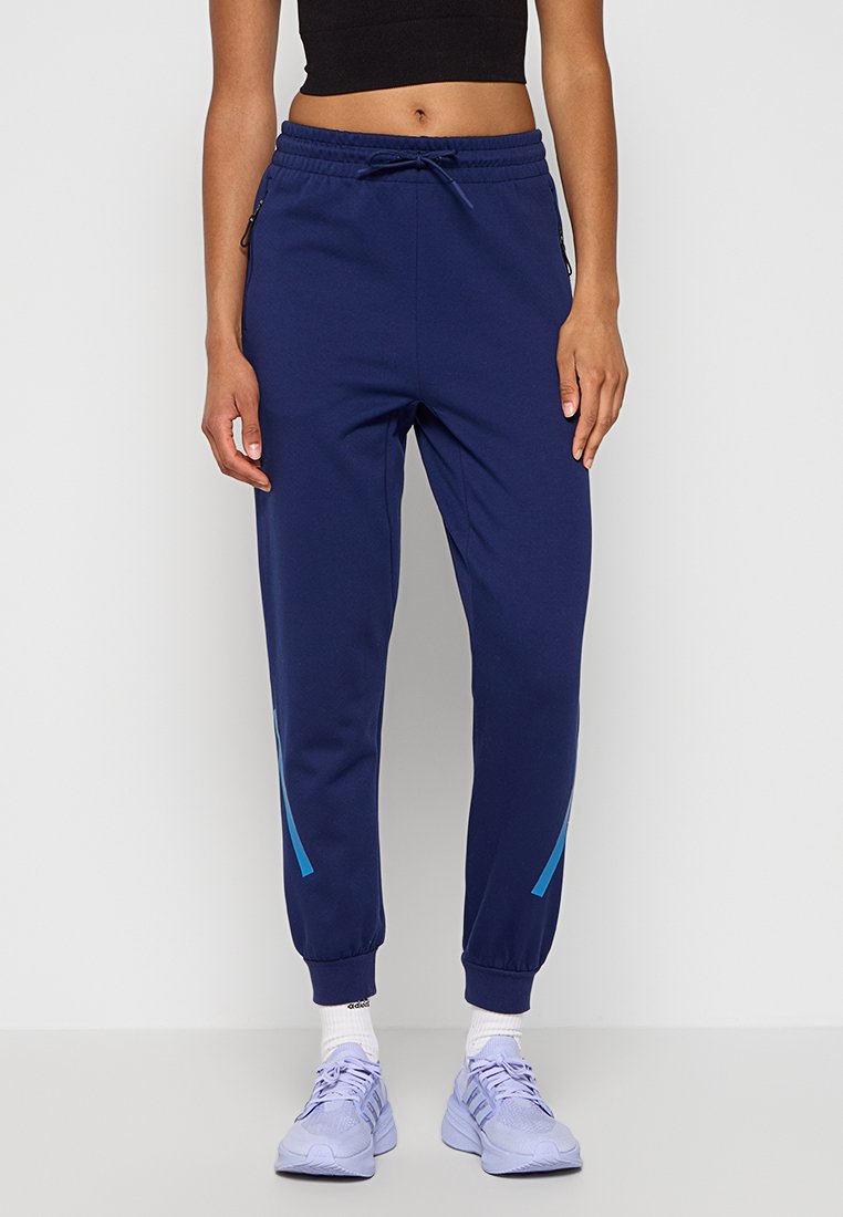 Person in dunkelblauen Jogginghosen mit hellblauen Streifen an den Knöcheln, schwarzem Crop-Top, weißen Socken und lila Laufschuhen, steht an einer weißen Wand.