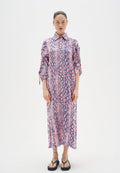Langes, rosa Schlangenmusterkleid mit kurzen Ärmeln, weitem Kragen und seitlichen Schlitzen. Getragen mit schwarzen Flip-Flops und minimalem Schmuck.