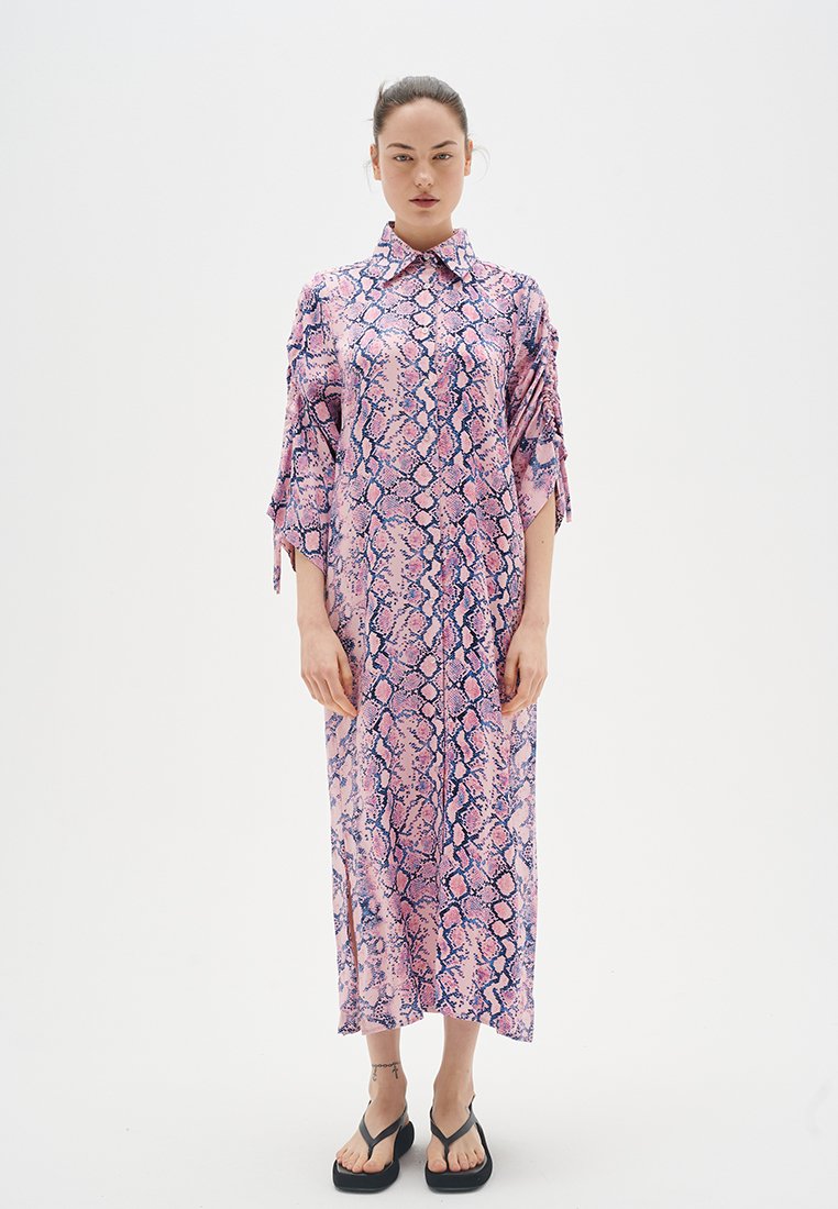 Langes, rosa Schlangenmusterkleid mit kurzen Ärmeln, weitem Kragen und seitlichen Schlitzen. Getragen mit schwarzen Flip-Flops und minimalem Schmuck.