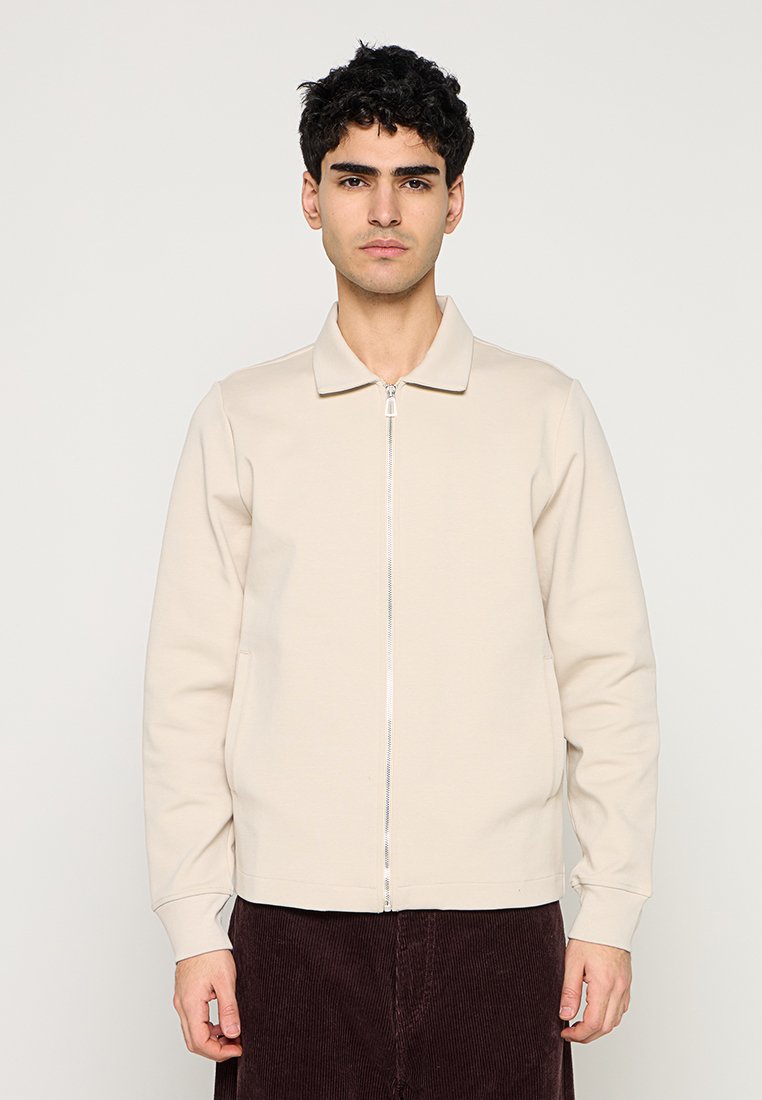 Beige Reißverschlussjacke aus weichem Stoff mit einem Kragen und gerippten Bündchen. Minimalistisches Design ohne sichtbare Muster oder Akzente.