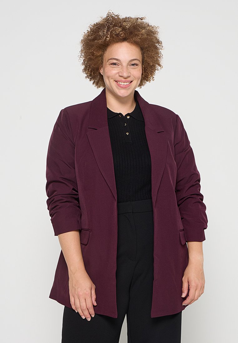 Burgunderfarbener Blazer mit lässiger Passform, langen Ärmeln und Fronttaschen, über einem schwarzen gerippten Oberteil und schwarzen Hosen getragen. Glatte Stofftextur.