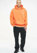 Oranger Hoodie mit vorderer Tasche, gerippten Bündchen und kleinem weißen Logo. Kombiniert mit schwarzen weiten Hosen und schwarzen Sneakers.