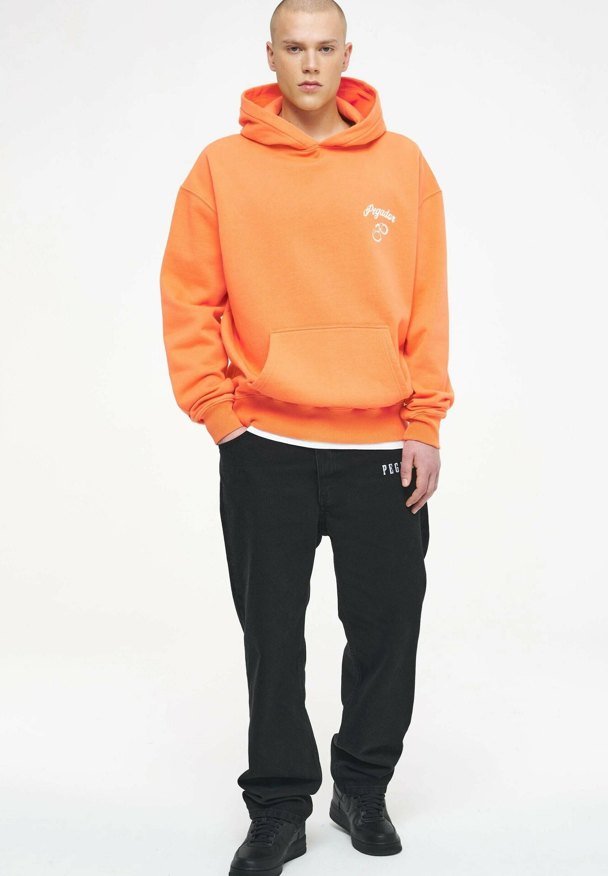 Oranger Hoodie mit vorderer Tasche, gerippten Bündchen und kleinem weißen Logo. Kombiniert mit schwarzen weiten Hosen und schwarzen Sneakers.