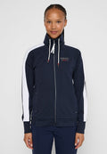 Junge Frau, die eine marineblaue Zip-Jacke mit weißen Streifen an den Ärmeln und dem Text "Sailing Crew" auf der Brust trägt.