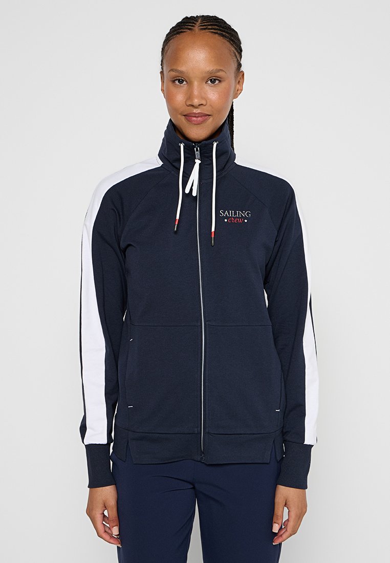 Junge Frau, die eine marineblaue Zip-Jacke mit weißen Streifen an den Ärmeln und dem Text "Sailing Crew" auf der Brust trägt.