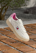 Weißer Ledersneaker mit rosa Innenfutter und beiger Sohle, mit einem kleinen Tommy Jeans-Logo an der Seite, auf Holzfliesen im Freien liegend.