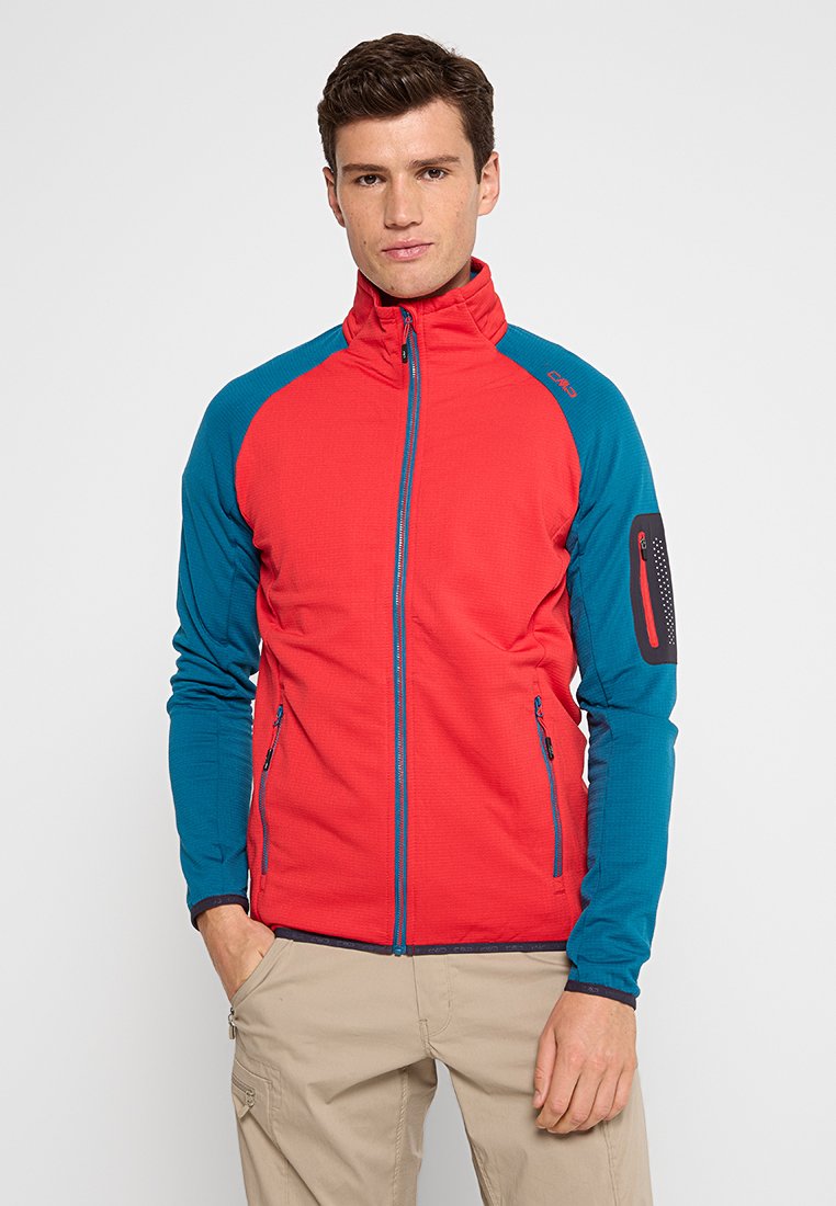 Mann trägt eine rot-blaue Jacke mit hohem Kragen und beigen Hosen, steht mit einer Hand in der Tasche vor einem weißen Hintergrund.
