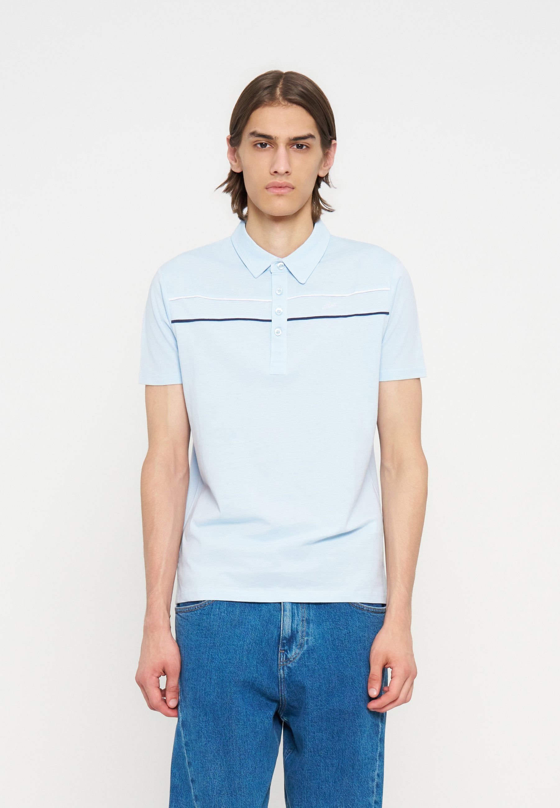 Junger Mann trägt ein hellblaues kurzärmeliges Poloshirt mit horizontalen Streifen, kombiniert mit blauen Jeans, steht vor einem einfarbigen Hintergrund.
