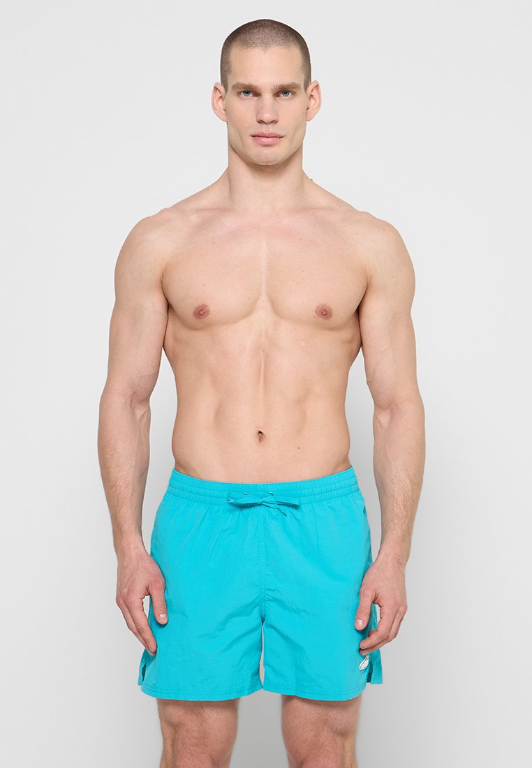 Muskelöser Mann steht nach vorne gerichtet und trägt leuchtend türkisfarbene Badeshorts mit Kordelzug an der Taille vor einfarbigem grauem Hintergrund.