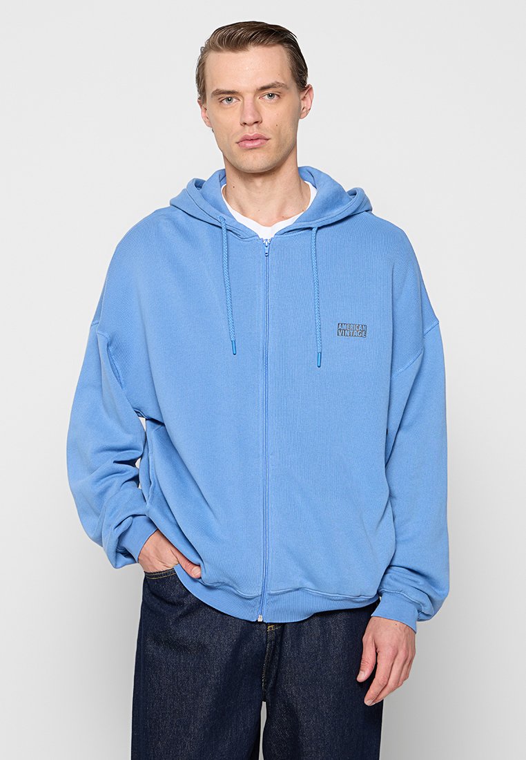 Hellblaues Zip-Hoodie aus strukturierter Stoff, mit einer Kordelzugkapuze und einem kleinen Logopatch auf der linken Brust. Lockere Passform.