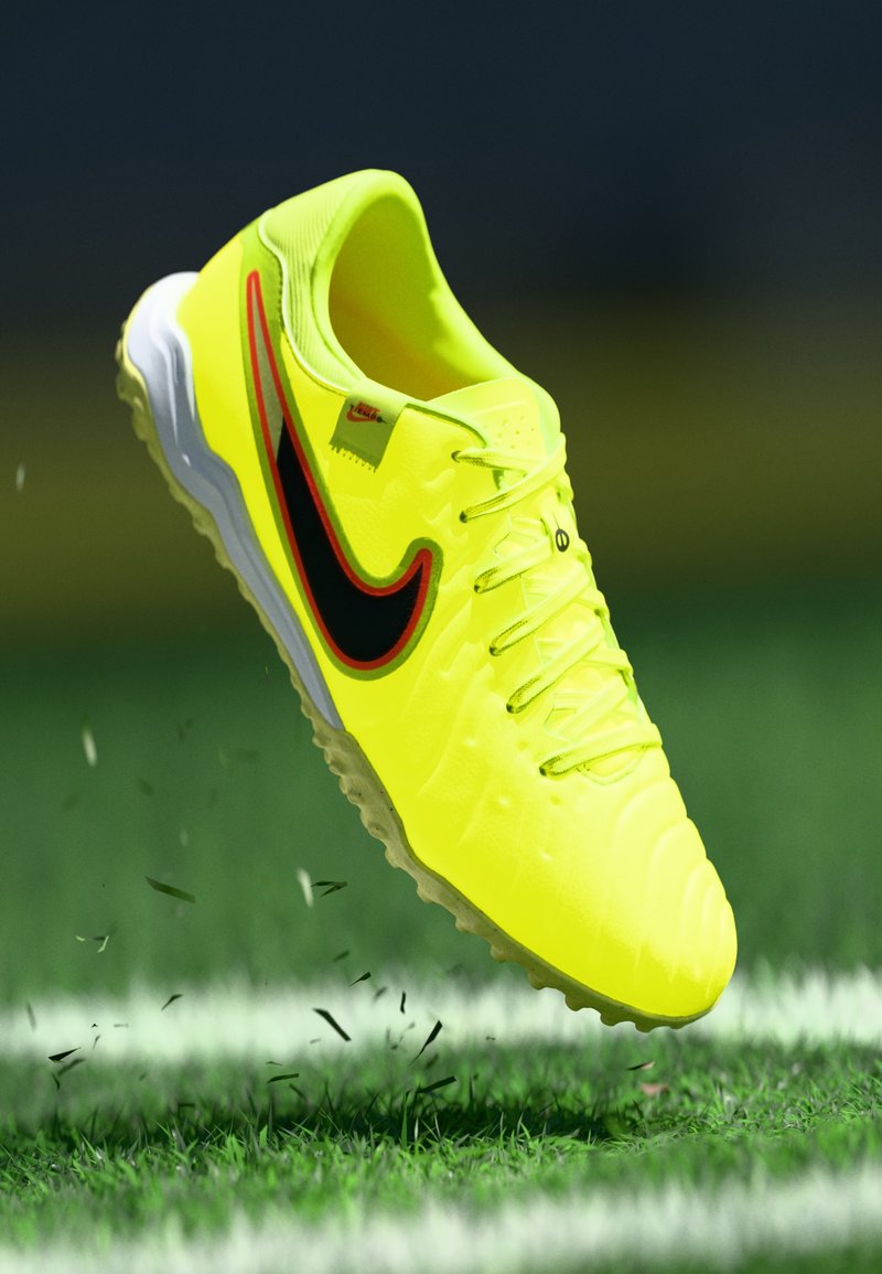 Helle gelbe Nike-Fußballschuhe mit schwarzem und rotem Swoosh, die über einem Rasenfeld mit weißen Linien und verstreuten Grasstücken schweben.