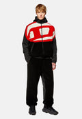 Männliches Outfit mit einer schwarzen Fleecejacke, die ein rotes und weißes abstraktes Design aufweist, und schwarzen Hosen mit einer strukturierten Oberfläche. Wuchtige schwarze Schuhe.