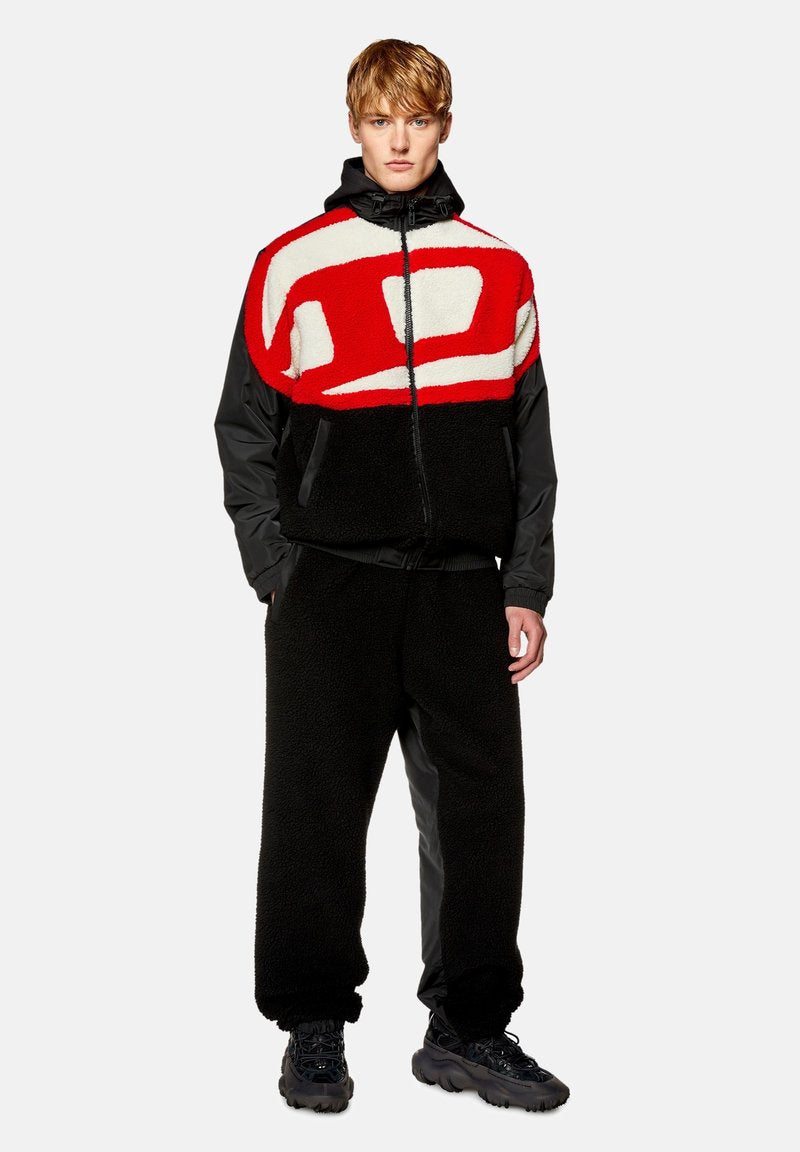 Männliches Outfit mit einer schwarzen Fleecejacke, die ein rotes und weißes abstraktes Design aufweist, und schwarzen Hosen mit einer strukturierten Oberfläche. Wuchtige schwarze Schuhe.