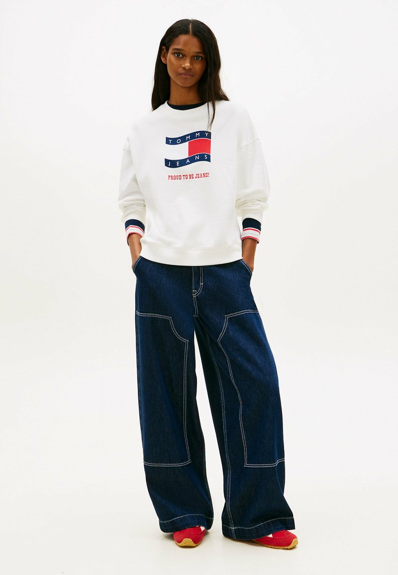 Frau mit langen Haaren, die ein weißes Tommy Jeans Sweatshirt, dunkle, weit geschnittene Jeans mit weißen Nähten und rote Sneaker trägt, stehend mit den Händen in den Taschen.
