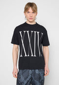 Junger Mann mit blondem Haar, der ein schwarzes T-Shirt mit großen weißen römischen Zahlen XXIX und gemusterten dunklen Shorts vor schlichtem Hintergrund trägt.