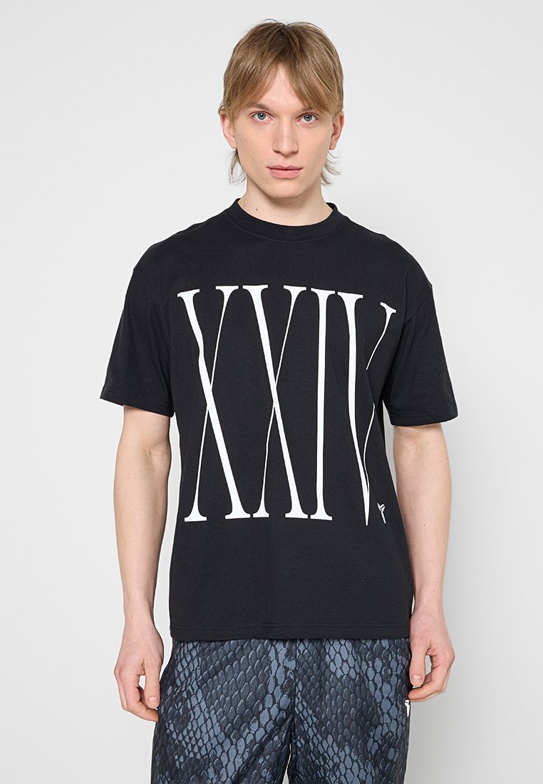 Junger Mann mit blondem Haar, der ein schwarzes T-Shirt mit großen weißen römischen Zahlen XXIX und gemusterten dunklen Shorts vor schlichtem Hintergrund trägt.