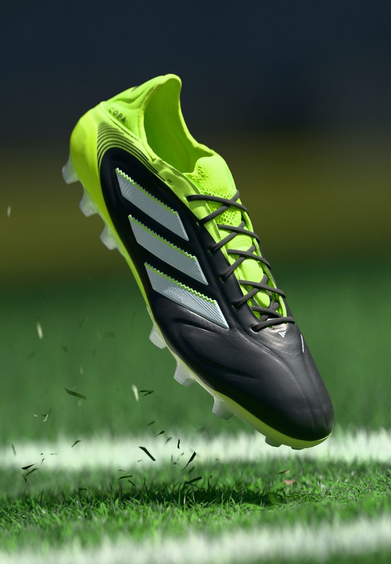 Schwarzer und neongelber Adidas-Fußballschuh in der Luft über Gras mit aufwirbelndem Dreck, der die Stollen und die drei weißen Streifen an der Seite zeigt.