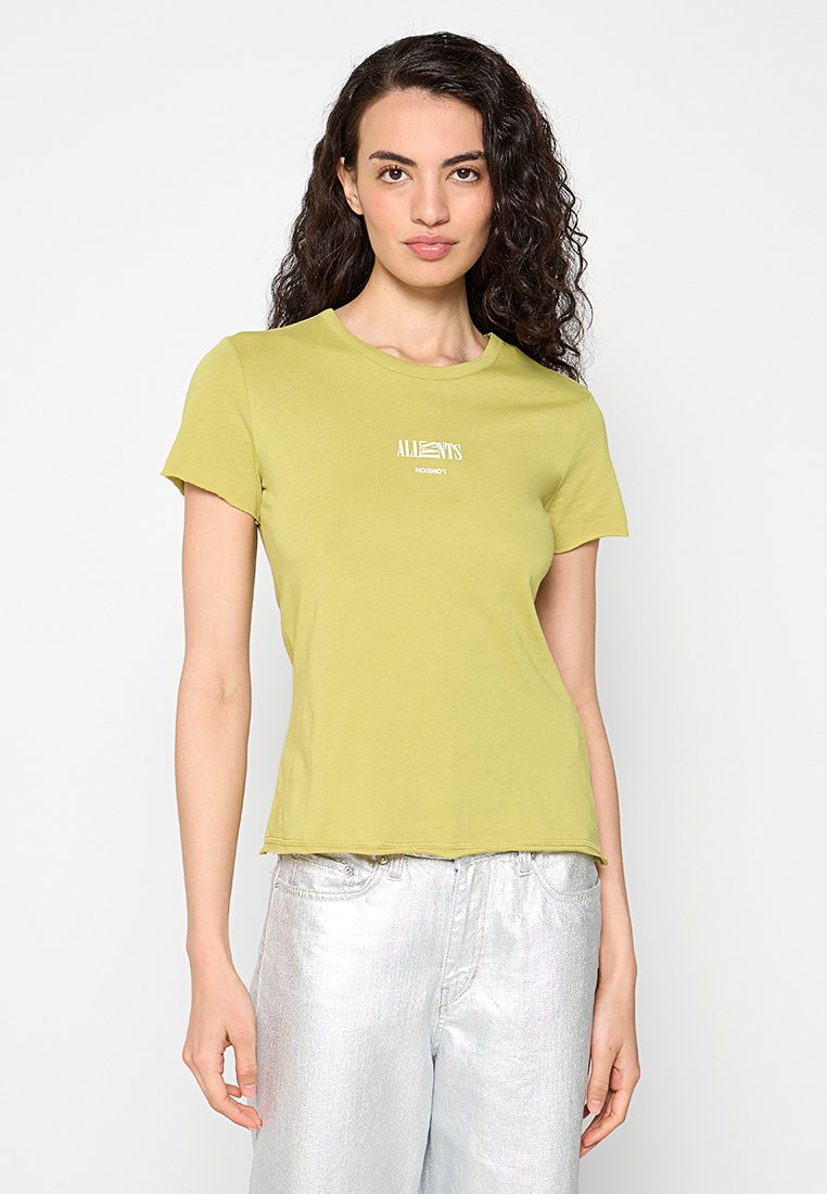 Frau mit lockigem dunklem Haar, die ein hellgrünes eng anliegendes T-Shirt mit kleinem weißem Text und glänzenden silbernen Hosen trägt, vor einem weißen Hintergrund stehend.