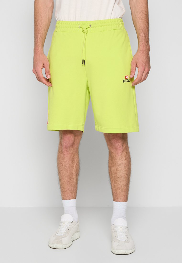 Mann in neon-gelben Diesel-Shorts mit Kordelzug, weißen Socken und weißen Turnschuhen, der vor einem schlichten Hintergrund steht.
