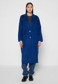 Langer blauer Wollmantel mit Reverskragen, goldener Knopfverschluss und zwei vorderen Taschen. Getragen über einem grauen Oberteil und kombiniert mit Jeans.