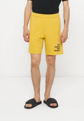 Mann trägt gelbe Shorts mit dem Text „The Future Surf Society“ am Bein, schwarzes T-Shirt und schwarze Slide-Sandalen, steht vor weißem Hintergrund.