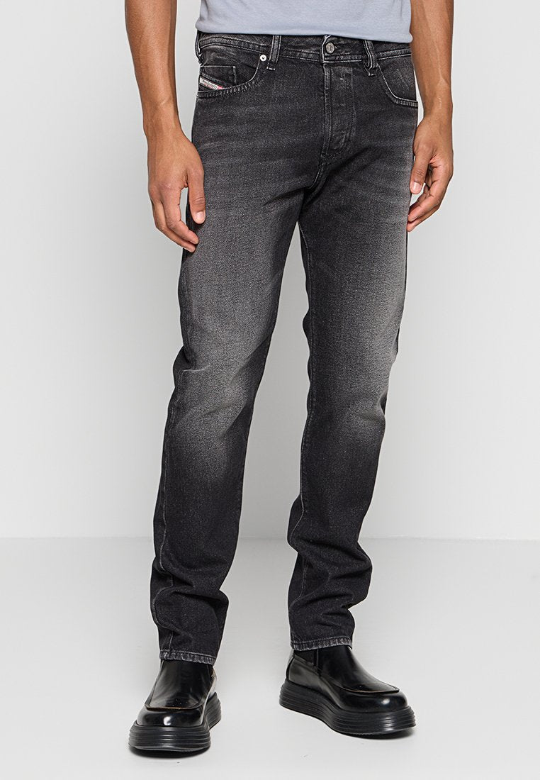 Mann trägt verwaschene schwarze Slim-Fit-Jeans und schwarze Plateauschuhe, steht vor einem schlichten grauen Hintergrund.