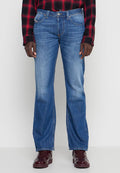 Mann trägt blaue Jeans mit geradem Bein, ein rot-schwarz kariertes Hemd und dunkelrote Turnschuhe, steht vor einem schlichten hellen Hintergrund.