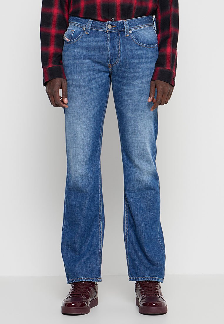 Mann trägt blaue Jeans mit geradem Bein, ein rot-schwarz kariertes Hemd und dunkelrote Turnschuhe, steht vor einem schlichten hellen Hintergrund.