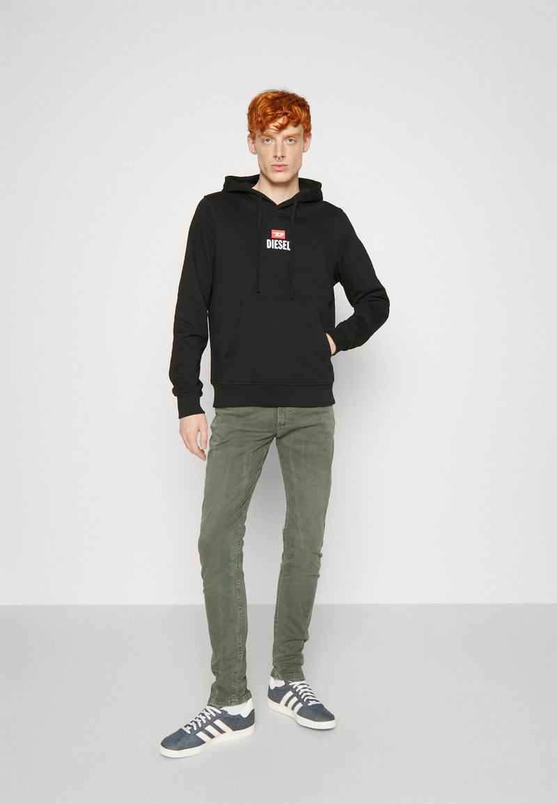 Schwarzer Kapuzenpullover mit Fronttasche und weißem "DIESEL"-Logo. Kombiniert mit olivgrünen Hosen und marineblauen Sneakers mit weißen Streifen.