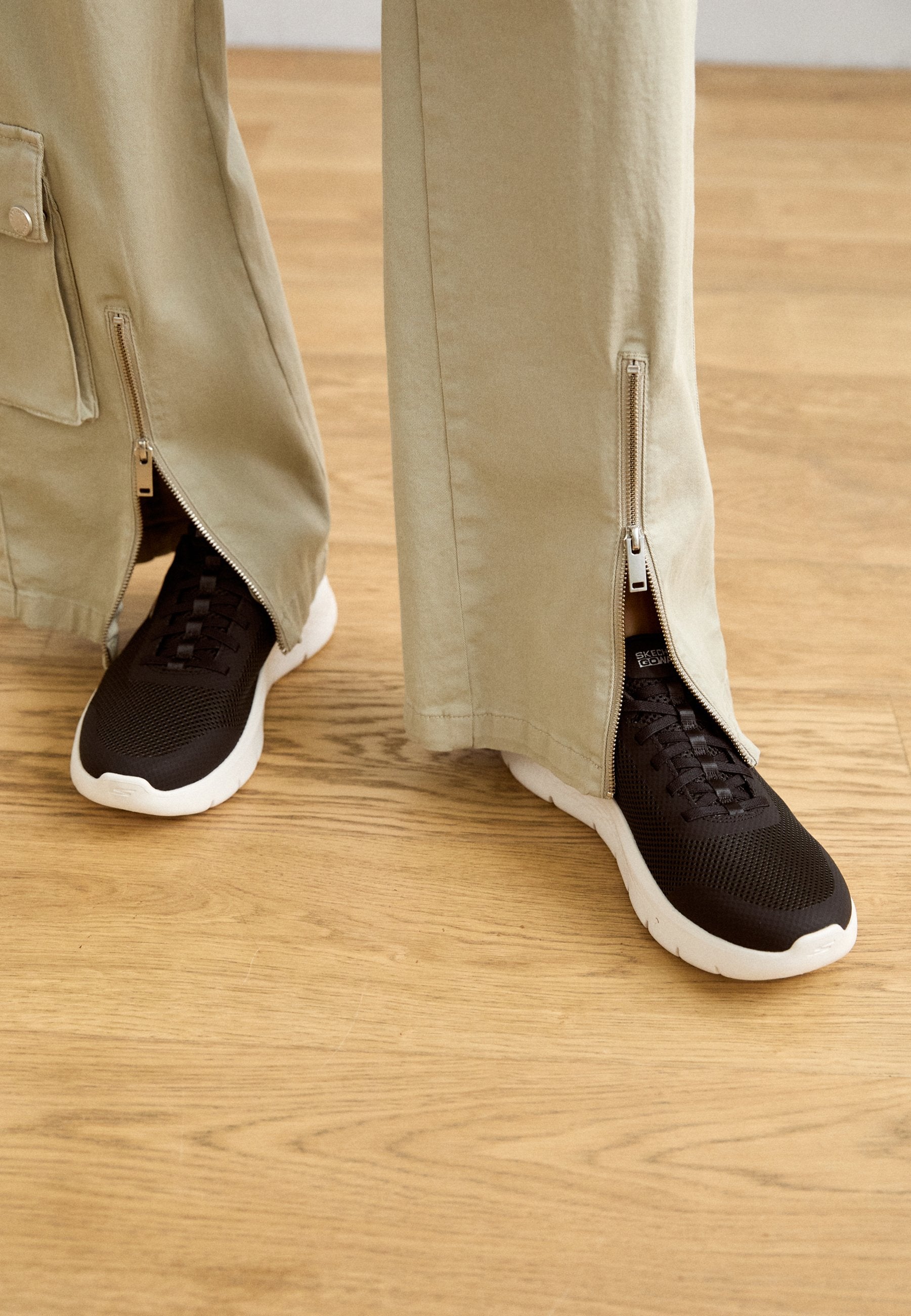 Schwarze strukturierte Sneaker mit weißen Sohlen sind teilweise von hellbeigen Hosen bedeckt, die seitliche Reißverschlüsse vom Saum bis zur Mitte haben.