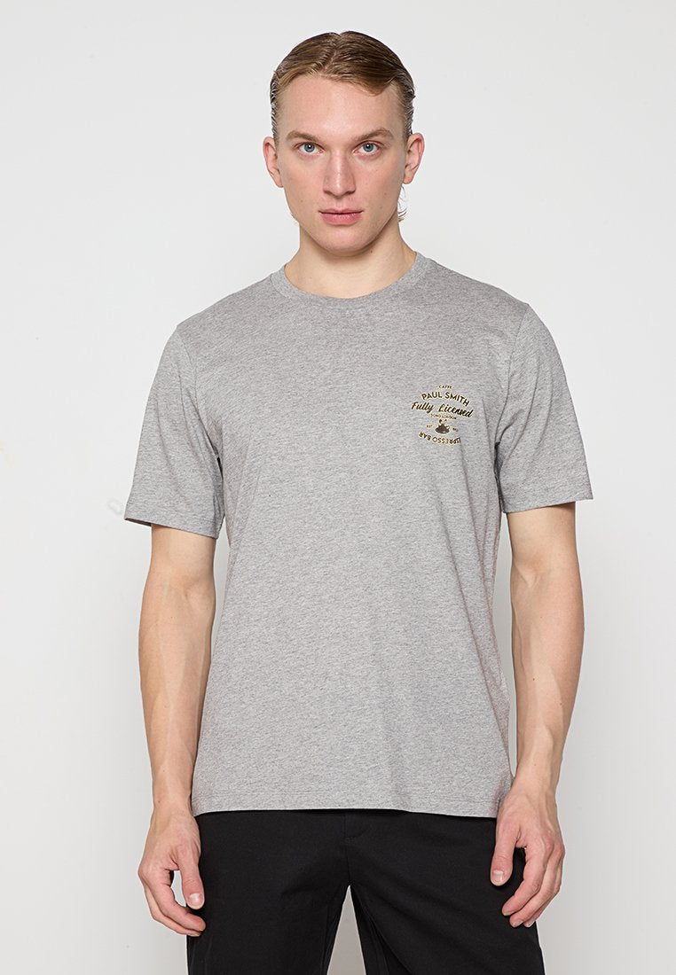 Junger Mann mit hellen Haaren, der ein graues Kurzarm-T-Shirt mit kleinem Text und Grafik auf der linken Brust und schwarze Hosen trägt.