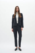 Dunkelgrauer, maßgeschneiderter Blazer und passende Hose, kombiniert mit einem weißen Rollkragenpullover. Die Schuhe sind spitze schwarze Ballerinas. Glattes Material und klare Linien.