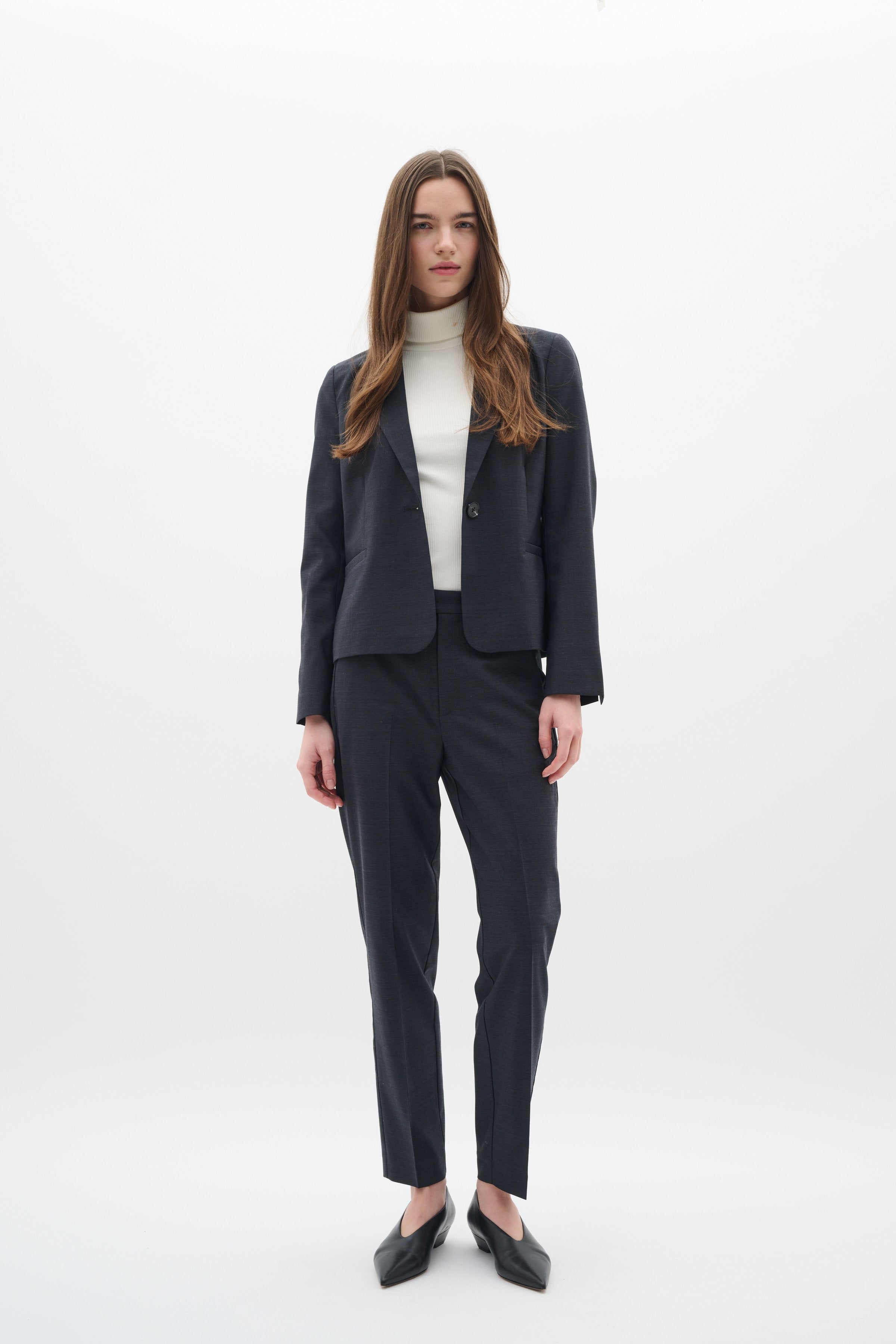 Dunkelgrauer, maßgeschneiderter Blazer und passende Hose, kombiniert mit einem weißen Rollkragenpullover. Die Schuhe sind spitze schwarze Ballerinas. Glattes Material und klare Linien.