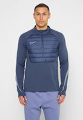 Mann trägt eine marineblaue Nike Steppjacke mit Viertel-Reißverschluss und reflektierenden Streifen an den Ärmeln sowie hellviolette Hose, steht vor weißem Hintergrund.