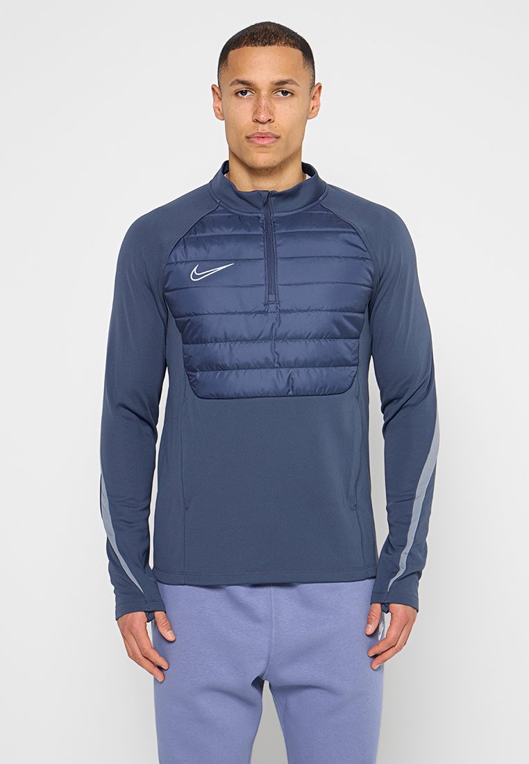 Mann trägt eine marineblaue Nike Steppjacke mit Viertel-Reißverschluss und reflektierenden Streifen an den Ärmeln sowie hellviolette Hose, steht vor weißem Hintergrund.