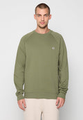 Mann mit kurzen Haaren, der einen olivgrünen Rundhalsausschnitt-Pullover mit langen Ärmeln und beigen Hosen trägt, steht vor einem schlichten hellen Hintergrund.