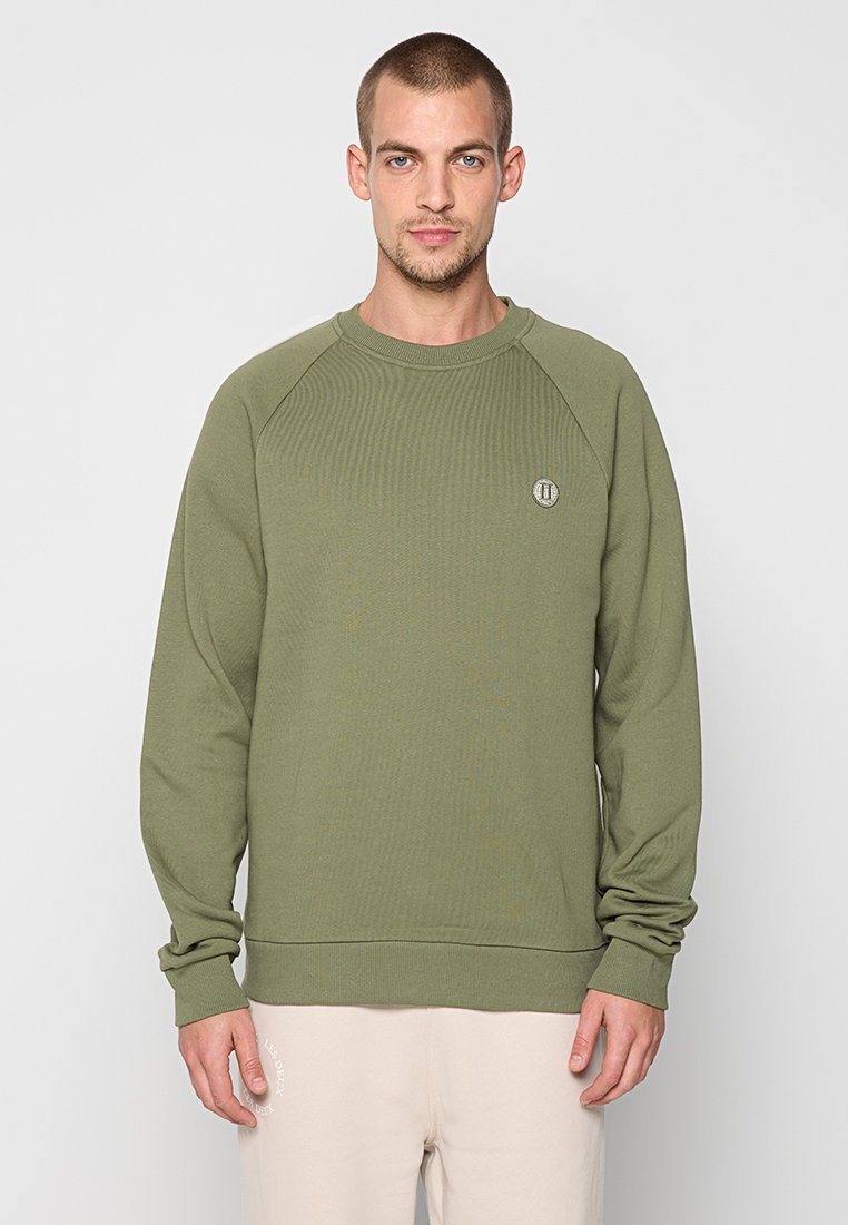 Mann mit kurzen Haaren, der einen olivgrünen Rundhalsausschnitt-Pullover mit langen Ärmeln und beigen Hosen trägt, steht vor einem schlichten hellen Hintergrund.