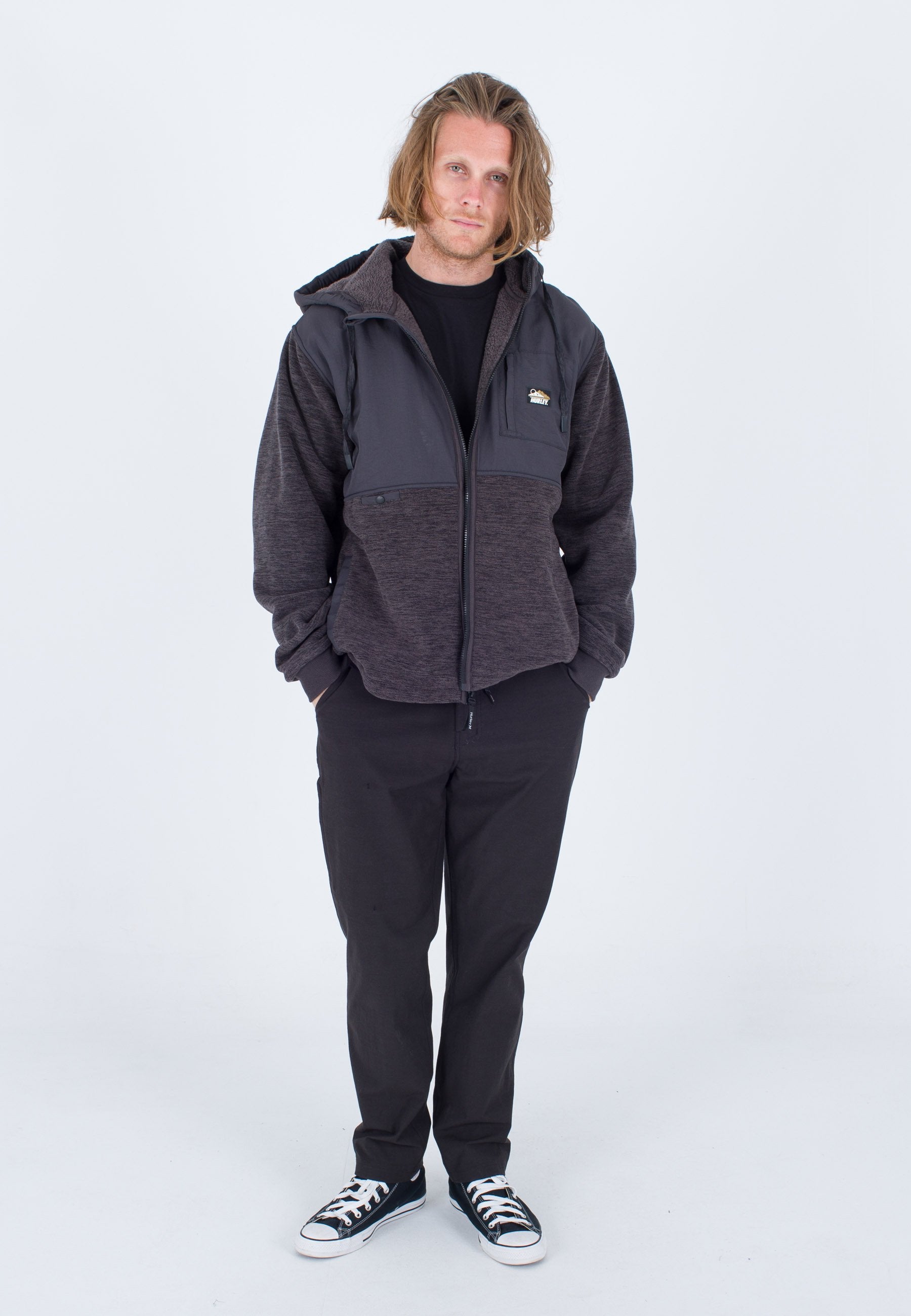 Grauer Fleecejacke mit Reißverschluß vorne, elastischen Bündchen und Seitentaschen; getragen mit einem schwarzen Hemd und dunklen Hosen. Schwarze Sneaker.