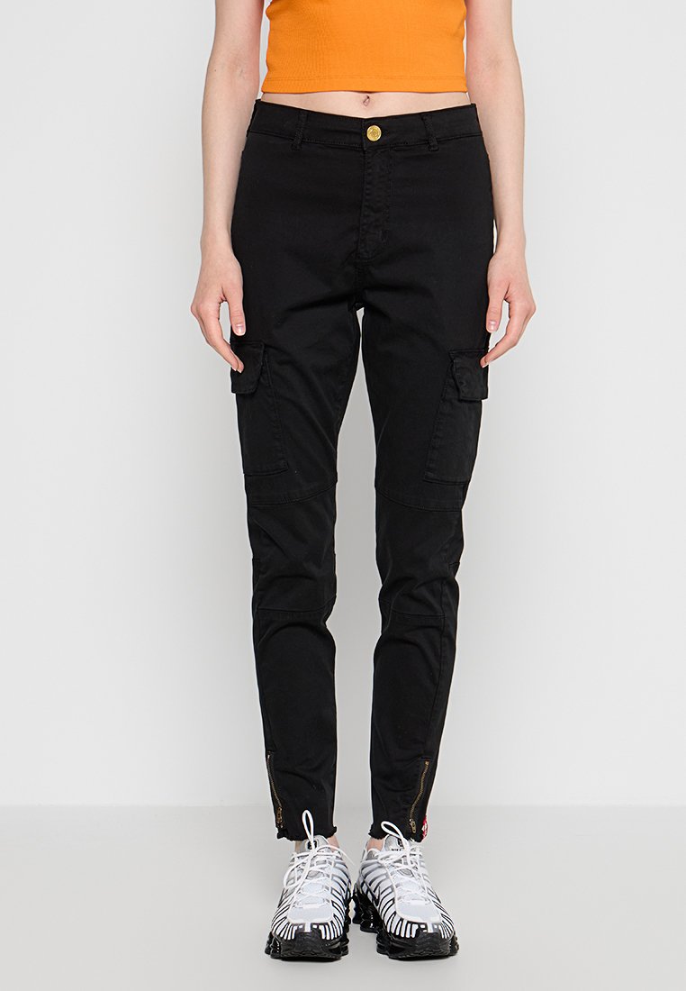 Person in schwarzen Cargo-Hosen mit Fronttaschen und weißen Sneakers, kombiniert mit einem orangefarbenen Cropped-Top, vor einem einfarbigen Hintergrund stehend.