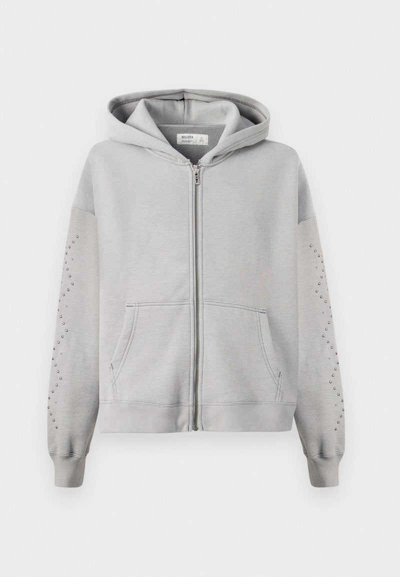 Grauer Kapuzensweatshirt mit einem durchgehenden Reißverschluss, das mit Nieten entlang der Ärmel und Kängurutaschen ausgestattet ist. Aus weichem Baumwollmaterial.