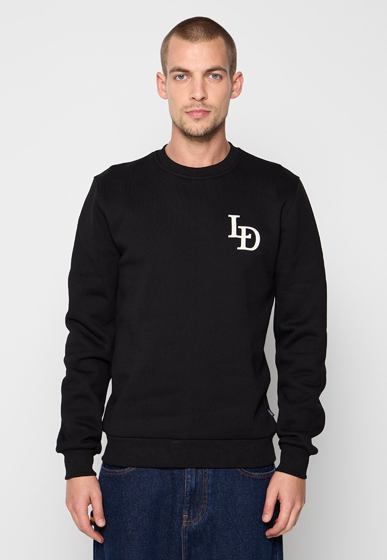 Junger Mann mit kurzen Haaren, der einen schwarzen Pullover mit "LD"-Logo auf der Brust und eine blaue Jeans trägt, steht vor einem schlichten hellen Hintergrund.