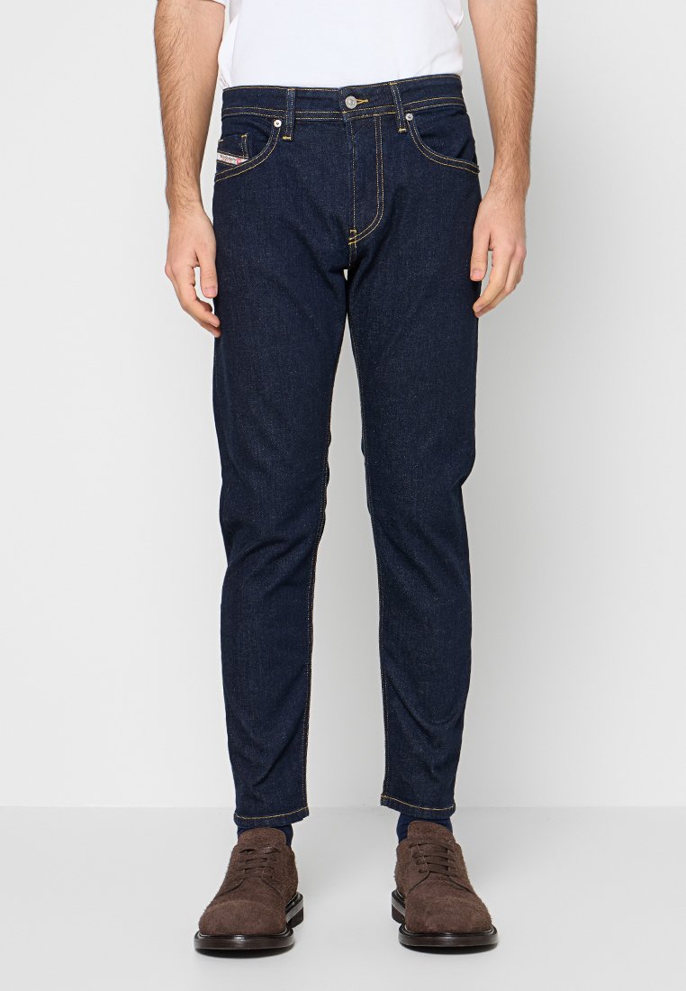 Dunkelblaue Jeans mit schmaler Passform, gelben Nähten, Fünf-Taschen-Design und Knopfverschluss. Kombiniert mit braunen Schuhen.
