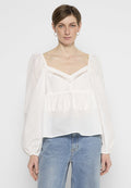 Weiße Bluse mit Ballonärmeln, V-Ausschnitt und dekorativem Fransen Detail entlang der Brust. Kombiniert mit blauen Denim-Jeans.